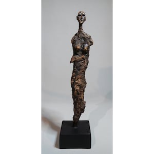May include: A bronze sculpture of a slender human figure with a textured surface. The figure is mounted on a black square base. The sculpture has a rough, organic appearance, with visible details suggesting a torso and head.