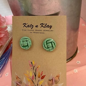 Pode incluir: Brincos de botão verdes com um design de nó, exibidos em um cartão castanho. O cartão apresenta o texto "Katz n Klay FINE HAND MADE JEWELRY IN MONTANA" e um desenho floral em aquarela. Os brincos são redondos e têm uma textura.