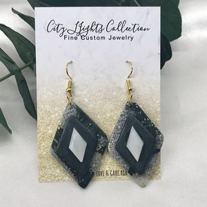 Może przedstawiać: Para kolczyków z wiszącym diamentowym wzorem. Kolczyki mają czarno-szarą nakrapianą podstawę, czarny kształt diamentu i białe diamentowe centrum. Złote haczyki i akcenty dopełniają wygląd. Na karcie napisano "City Lights Collection Fine Custom Jewelry."