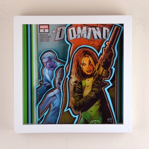 May include: A framed comic book cover featuring Domino, a Marvel Comics character, holding a gun. The cover is in a white frame with a green and blue border. The title "DOMINO" is printed in large letters across the top of the cover.