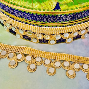 May include: Gold trim with round mirror embellishments. The trim is attached to a white fabric background.