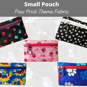 May include: Small pouches with paw print theme fabric. The pouches have zippers and small metal charms. The fabrics are black, white, pink, blue, and tie-dye, with colorful paw prints and bone designs. The text "Small Pouch Paw Print Theme Fabric" is at the top.