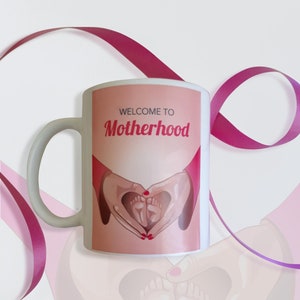 Puede incluir: Taza de cerámica blanca con una ilustración rosa y roja de manos formando un corazón alrededor de dos pies de bebé. El texto "Bienvenido a la maternidad" está impreso en negro en la taza.