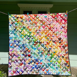 May include: A colorful handmade quilt with a vibrant patchwork design. The quilt features a diagonal pattern of squares in a rainbow of colors, including red, orange, yellow, green, blue, and purple. It is hanging on a clothesline outdoors.