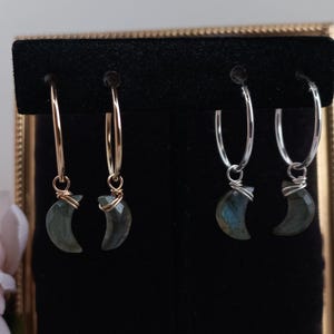 May include: A pair of hoop earrings with crescent moon shaped labradorite stones hanging from them. The hoops are available in gold and silver.