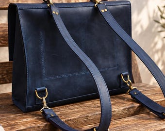 May include: A dark blue leather backpack with a rectangular shape, featuring a top handle and two adjustable shoulder straps attached with brass-toned metal hardware. The bag has a flap closure and visible stitching details on the back panel.