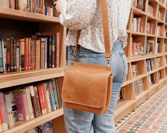 Könnte beinhalten: Eine Frau in einer weißen Blusen mit Blumenmuster und blauen Jeans stöbert in den Regalen einer Buchhandlung. Sie trägt eine braune Ledertasche mit einem langen Riemen.