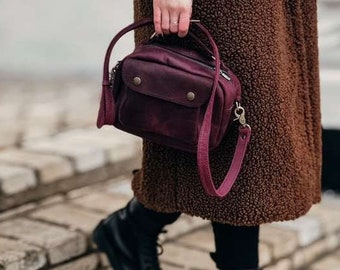 May include: A burgundy leather satchel bag with a flap closure and a long strap. The bag is worn by a person wearing black boots and a brown coat.