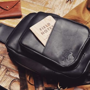 May include: A black leather crossbody bag with a zipper closure and a small pocket on the front. The pocket has a wooden card inside that says "Field Notes".