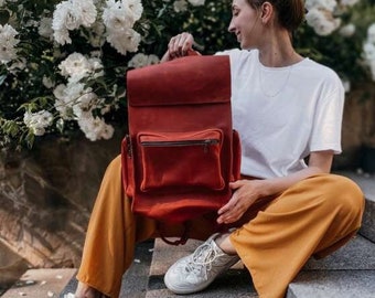 Può includere: Uno zaino in pelle rossa con una tasca con cerniera sul davanti. Lo zaino è portato da una persona che indossa una maglietta bianca e pantaloni gialli.