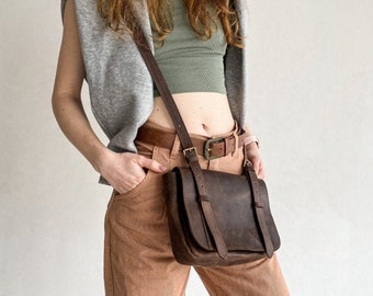 Könnte beinhalten: Eine braune Leder-Umhängetasche mit einer Klappe und einer Schnalle. Die Tasche wird über einem grauen ärmellosen Oberteil und einer braunen Hose getragen.