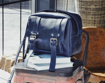 May include: A dark blue leather backpack with two adjustable straps and two brass buckles. The backpack is sitting on a wooden chair with a stack of books.