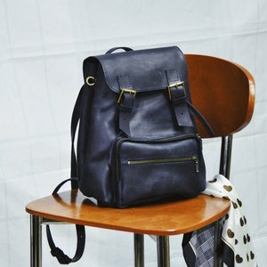 May include: A dark blue leather backpack with brass buckles and a zippered front pocket. The backpack is sitting on a wooden chair.
