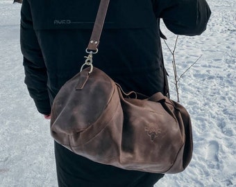 以下が含まれることがあります： 肩にかけられた黒いジャケットを着た人の肩にかけられた、ストラップと金色のバックルが付いた茶色の革製のダッフルバッグ。