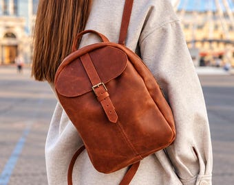 May include: A medium-sized, cognac-colored leather backpack featuring a fold-over flap with a vertical strap and a metal buckle closure. The backpack has a top handle and two adjustable shoulder straps, showcasing a smooth, matte finish.