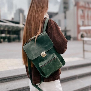 May include: A dark green leather backpack with a flap closure and a zippered pocket. The backpack has a brass buckle and a strap.