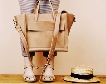 May include: A tan leather tote bag with a long strap and a smaller pocket on the front. The bag has the word "ARTIGIAL" printed on the strap. The bag is being held by a person wearing a patterned skirt and sandals.