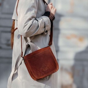 May include: A brown leather crossbody bag with a flap closure and a silver buckle. The bag has a long strap and is worn over the shoulder.