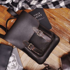 May include: A dark brown leather backpack featuring a top flap secured by two buckled straps, a top handle, and adjustable shoulder straps. The backpack is positioned alongside a Field Notes notebook, a bottle, a wallet, and a stringed instrument.