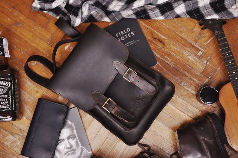 May include: A dark brown leather backpack featuring a top flap secured by two buckled straps, a top handle, and adjustable shoulder straps. The backpack is positioned alongside a Field Notes notebook, a bottle, a wallet, and a stringed instrument.