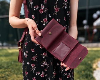 May include: A deep burgundy leather wallet featuring a tri-fold design, a clear ID window, multiple card slots, and a brass-colored snap button closure. The wallet is held open to display its interior compartments and stitching details.