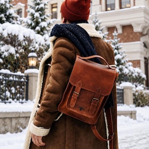 May include: A brown leather backpack with a top handle, a fold-over flap, and two vertical straps with metal buckles. The backpack features a small embossed bull logo on the front. It is worn over a brown suede coat with a white shearling lining.