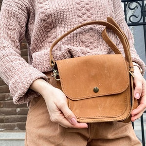 May include: A brown leather crossbody bag with a flap closure and a single strap. The bag has a small pocket on the front and a metal clasp.