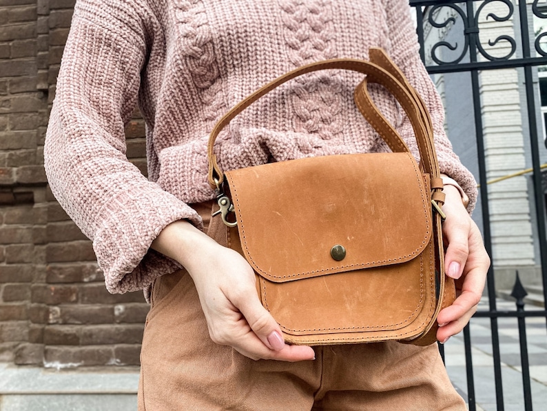 May include: A brown leather crossbody bag with a flap closure and a single strap. The bag has a small pocket on the front and a metal clasp.