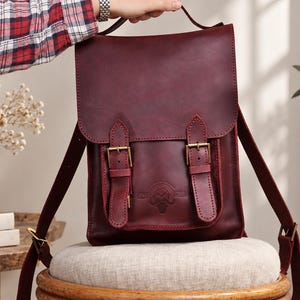 May include: A deep burgundy leather backpack featuring a fold-over flap with two vertical straps and brass-toned metal buckles. The front pocket displays a circular embossed logo. The bag includes adjustable shoulder straps and a top handle.