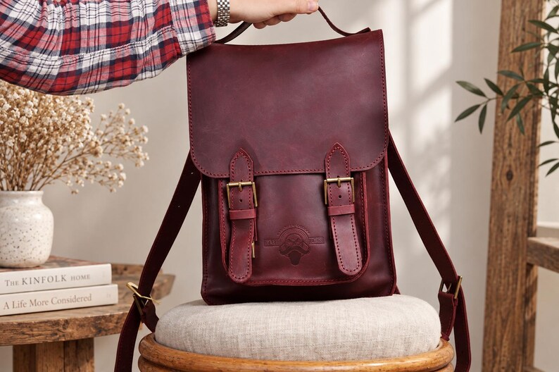 May include: A deep burgundy leather backpack featuring a fold-over flap with two vertical straps and brass-toned metal buckles. The front pocket displays a circular embossed logo. The bag includes adjustable shoulder straps and a top handle.