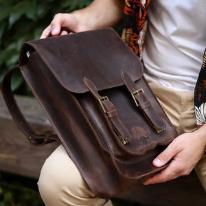 May include: A dark brown leather backpack featuring a fold-over flap, two vertical straps with brass-toned metal buckles, and a front pocket. The bag has a smooth, matte texture and visible stitching along the edges of the flap and the front pocket.