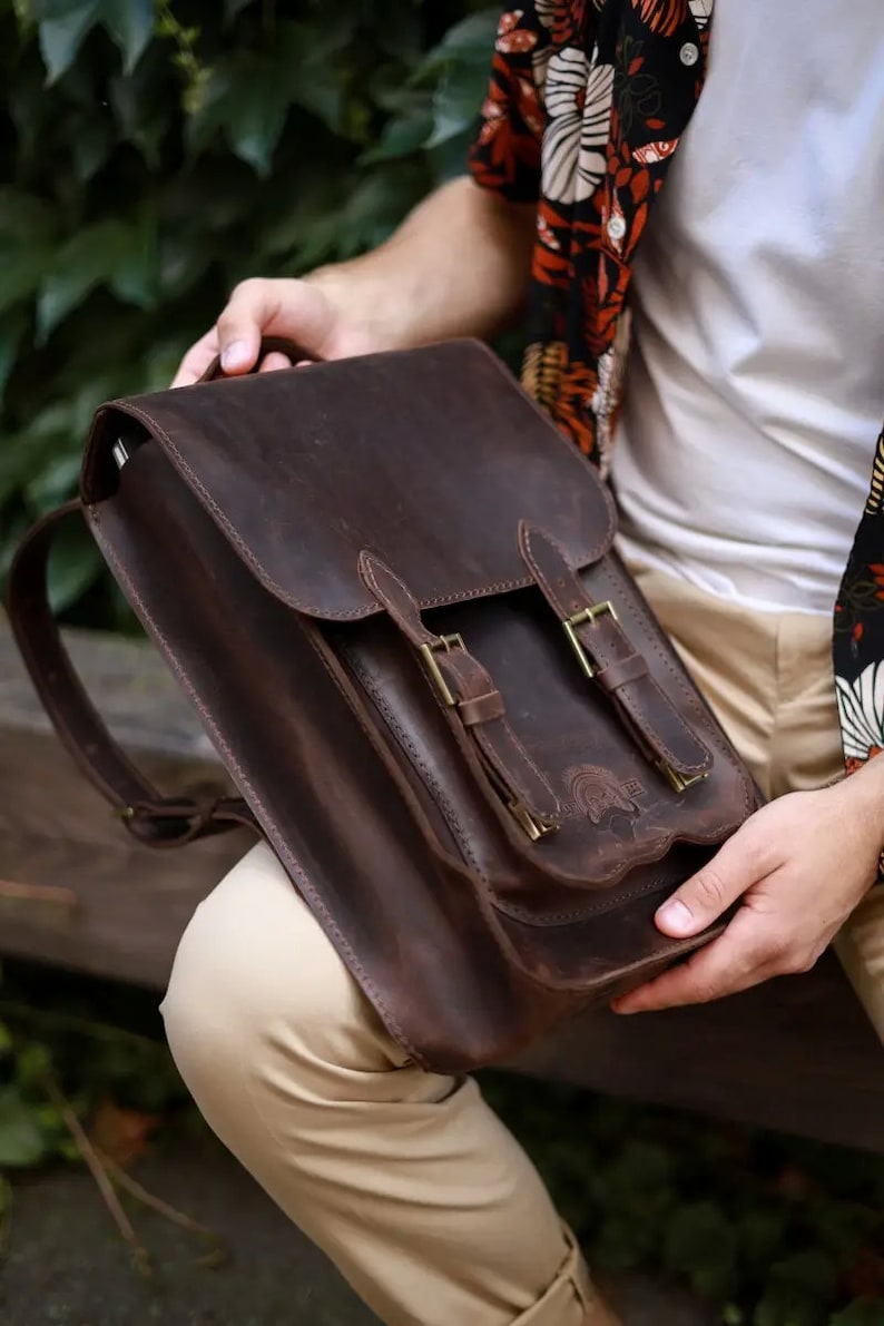 May include: A dark brown leather backpack featuring a fold-over flap, two vertical straps with brass-toned metal buckles, and a front pocket. The bag has a smooth, matte texture and visible stitching along the edges of the flap and the front pocket.