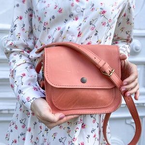 May include: A coral pink leather crossbody bag with a flap closure and a gold buckle. The bag has a long adjustable strap and is worn by a person wearing a white floral dress.