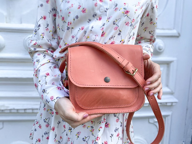 May include: A coral pink leather crossbody bag with a flap closure and a gold buckle. The bag has a long adjustable strap and is worn by a person wearing a white floral dress.
