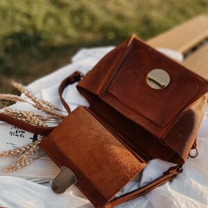 May include: A brown leather crossbody bag with a flap closure and a circular metal accent. The bag has a long strap and a smaller pocket on the front.