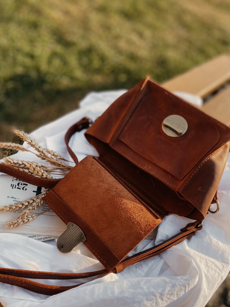 May include: A brown leather crossbody bag with a flap closure and a circular metal accent. The bag has a long strap and a smaller pocket on the front.