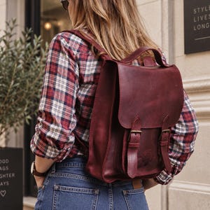 May include: A deep burgundy leather backpack featuring a top handle, a fold-over flap with two vertical straps, and antique brass-toned metal buckles. The backpack is worn over a red, white, and blue plaid long-sleeve shirt and blue denim jeans.
