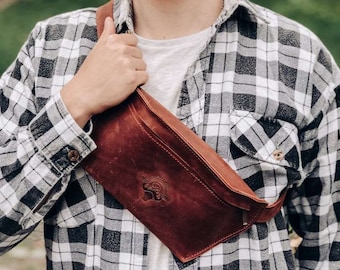 May include: A brown leather fanny pack with a flap closure and adjustable strap. The bag features a stamped design on the front and is worn across the body. The person is wearing a black and white plaid shirt.