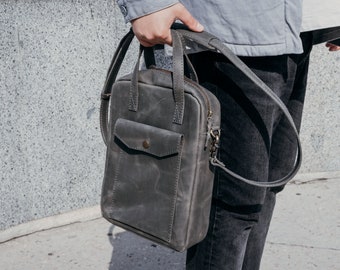 May include: A gray leather messenger bag with a zipper closure and a front pocket. The bag has a long adjustable strap.