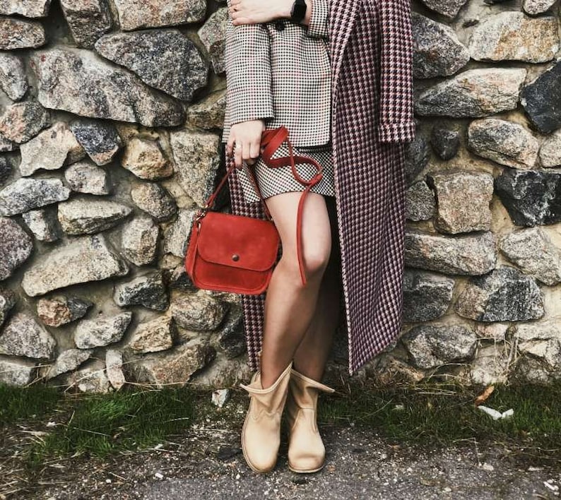 May include: A woman wearing a red and white checkered coat and a red leather shoulder bag. She is wearing tan ankle boots and standing in front of a stone wall.