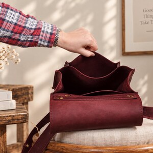 May include: A deep burgundy leather bag with an open top, featuring a structured rectangular base, a smooth matte finish, and a matching leather strap. The interior is lined with the same dark red material, showing a clean, minimalist construction.