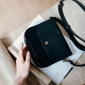 May include: A black leather crossbody bag with a flap closure and a gold button. The bag has a long adjustable strap.