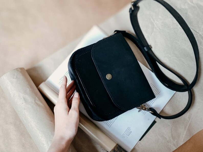 May include: A black leather crossbody bag with a flap closure and a gold button. The bag has a long adjustable strap.