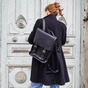 May include: A black leather backpack with a fold-over flap, two vertical straps with metal buckles, and adjustable shoulder straps. The bag is worn over a dark coat, featuring a structured rectangular shape with visible stitching and smooth leather.