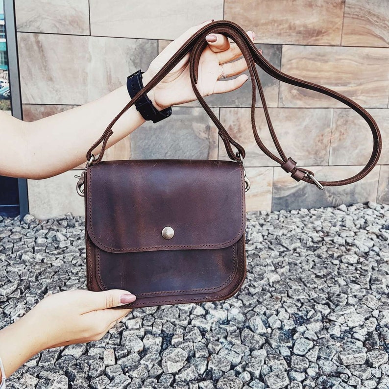 May include: A brown leather crossbody bag with a flap closure and a silver button. The bag has a long adjustable strap.