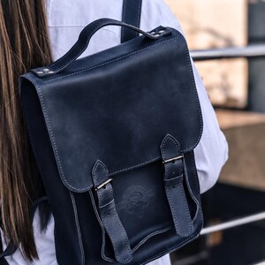 May include: A dark navy blue leather backpack featuring a top handle, a fold-over flap with two vertical buckled straps, and a circular embossed logo on the front. The bag has visible stitching along the edges and is worn on the back.