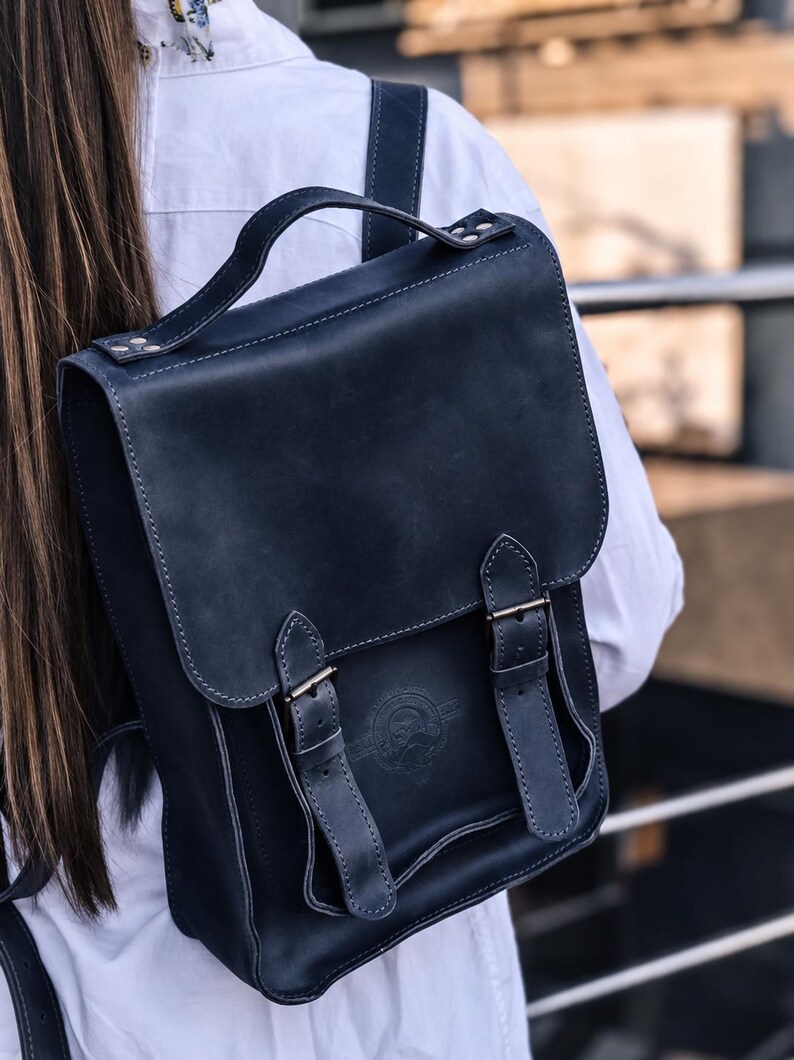 May include: A dark navy blue leather backpack featuring a top handle, a fold-over flap with two vertical buckled straps, and a circular embossed logo on the front. The bag has visible stitching along the edges and is worn on the back.