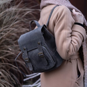 May include: A grey leather backpack with two adjustable buckles and a zipped pocket. The backpack is being worn by a person wearing a beige coat and a scarf.