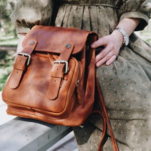 Peut inclure: Un sac à dos en cuir marron avec deux grandes boucles sur le devant. Le sac à dos a une poche zippée sur le devant et une plus petite poche sur le côté.