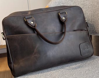 Pode incluir: Uma pasta de couro castanho escuro com fecho de correr superior, duas alças e um bolso frontal. A mala tem ferragens douradas e um pequeno emblema em relevo no canto inferior direito. A pasta está numa cadeira cinzenta.